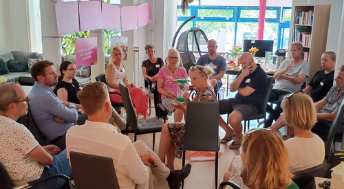 15 Menschen sitzen in zwei Stuhlkreisen. Frau mit mittleren Kreis hält eine grüne Sanduhr durch die grüner Sand rinnt.