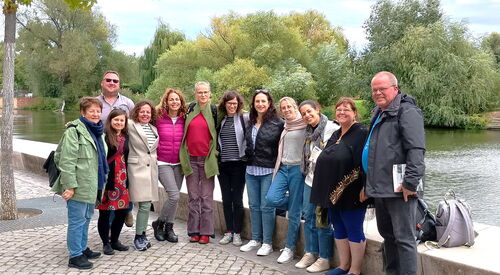 Gruppenbild. 10 Frauen, 2 Männer stehen an einem Fluss. Bäume und viel grün im Hintergrund auf der anderen Flussseite.