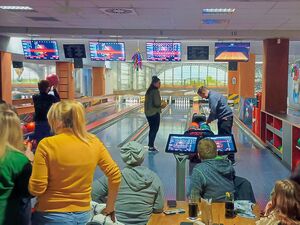 Eine Bowlinghalle. Es wird gebowlt, einige sitzen am Tisch mit Getränken und schauen zu.