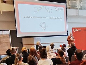 Präsentation an der Leinwand vorn im Raum. Viele Teilnehmende auf Stühlen.