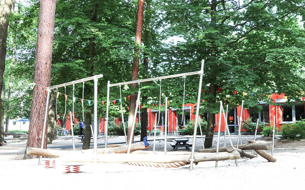 Spielplatz zwischen Bäumen. Durch die Bäume erkennt man das rote Kita Gebäude.