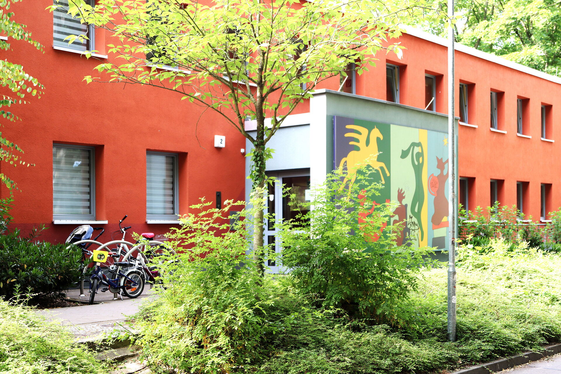 Zweistöckiges rotes Haus, Büsche und kleine Bäume davor. Der Eingangsbereich ist bunt gestaltet, an der Wand sind Bilder, ein geflügeltes Pferd, eine Tänzerin.