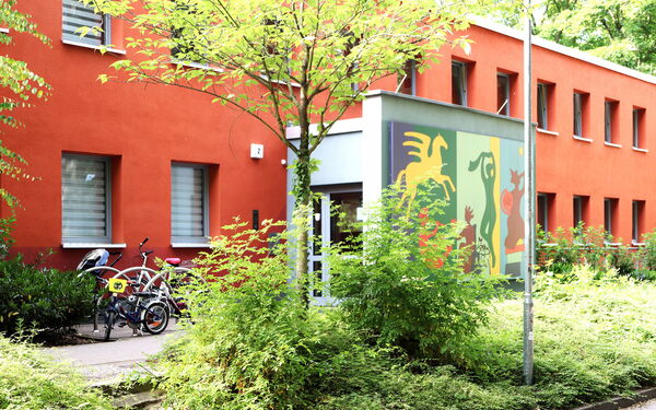 Zweistöckiges rotes Haus, Büsche und kleine Bäume davor. Der Eingangsbereich ist bunt gestaltet, an der Wand sind Bilder, ein geflügeltes Pferd, eine Tänzerin.