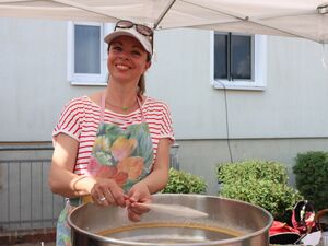 Eine junge Frau steht an einer Maschine zum Zuckerwatte machen und dreht ein Holzstäbchen um das sich die Zuckerwatte wickelt. Die Frau tragt ein T-Shirt, eine Schürze und ein Basecap und lächelt.