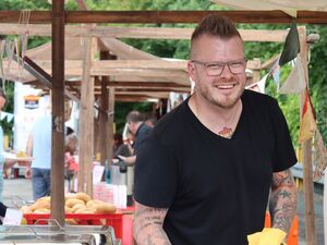 Mann an einem Stand lächelt fröhlich in die Kamera. Er trägt eine UnderCut Frisur, eine Brille und ein schwarzes T-Shirt. Er hat viele Tattoos.