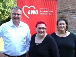 Portrait: ein Mann und zwei Frauen stehen draußen vor einem roten AWO Roll-Up-Banner. Mann in weißem Hemd, Frau mit roten kurzen Haare, roter Brille und roten Lippen und Frau mit langen dunklen Haaren, Ohrringen und Kette.