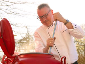 Koch mit weißer Kochjacke und Brille steht an einem großen roten Kessel und rührt um.