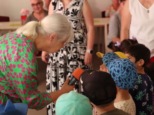 Kita Kinder werden von Frau mit Mikro interviewt. Frau hat graue Haare und ein grünes Kleid mit pinken Tupfen. Die Kinder tragen Basecaps.