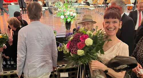 Eine Frau mit einem großen Blumenstrauß in heller Bluse sowie Hose. Sie steht in einer Sitzreihe des Nikolaisaals uf der Bühne sind noch Musiker.