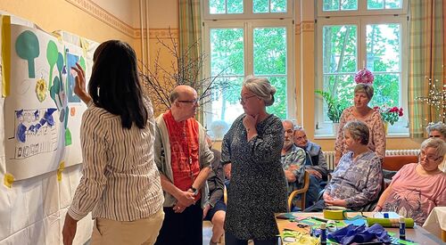 Ein Raum, große Fenster. Ältere Menschen sitzen auf Stühlen. Zwei ältere Menschen reden vor miteinander. Eine Frau mit langen dunklen Haaren gestaltet ein Plakat, diese Frau ist von hinten fotografiert.