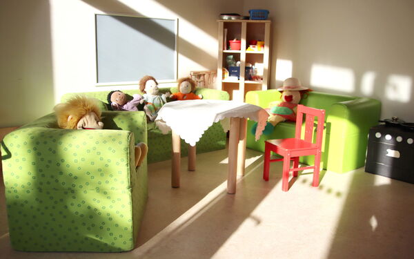 Stoffpuppen auf zwei grünen Kindersofas und einem Sessel. Ein roter Kinderstuhl steht an einem kleinen Tisch mit weißem Tischtuch in der Mitte.