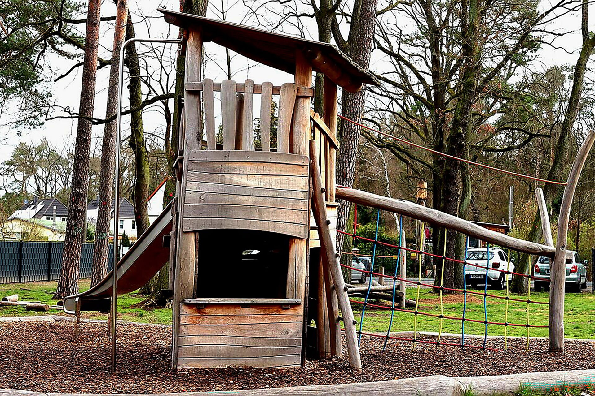 Spielplatz: Ein Holzspielturm mit einer Rutsche und einer kleinen Plattform, umgeben von Bäumen.