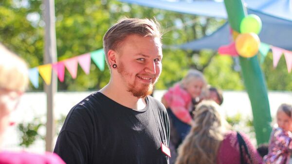 Ein Erzieher lächlet in die Kamera. Junger Mann mit kurzen braunen Haaren und Bart, schwarzes T-Shirt. Draußen, die Sonne scheint. Es sind bunte Wimpel und Luftballons aufgehängt.