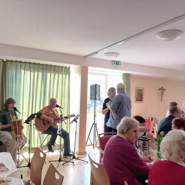 Heller Raum mit großen Fenstern. Kaffeetafeln und zwei Menschen die am Fenster sitzen und Musik machen.