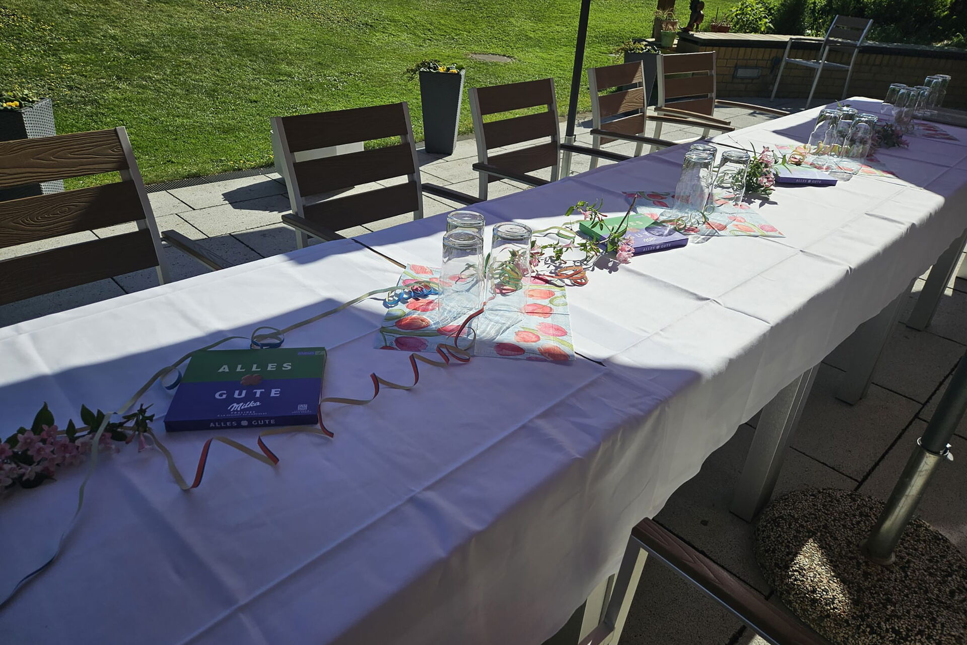 Lange Kaffeetafel auf der Terrasse, geschmückt mit Luftschlagen. Milka Schokoladenpackungen mit "Alles Gute" liegen auf dem weißen Tischtuch.
