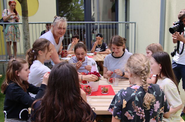 Draußen vor einem Gebäude. Kinder sitzen an Tischen und essen gut gelaunt Erdbeeren. Eine blonde Frau mit blonden Haaren und Brille auf den Kopf geschoben, steht am vorderen Tisch und lächelt.