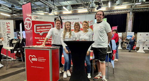 AWO Potsdam Messestand in einer Halle. 5 Mitarbeitende 3 Frauen eingerahmt von zwei Männern sie stehen hinter einem schwarzen Stehtisch. Messetheke mit AWO Logo Druck.