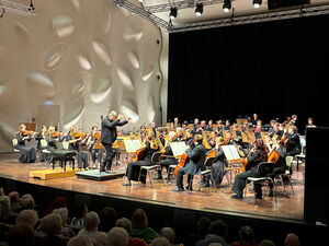 Große Bühne, großes Orchester. Dirigent vorne auf eine Plattform mit schwungvollen Armbewegungen.