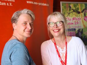 Zwei Frauen. Eine mit kurzen Haaren, die andere hat blonde lange Haare und eine große runde Brille mit dunklem Rahmen. Im Hintergrund das Plakat vom Theaterstück: Die Reise zum Ich