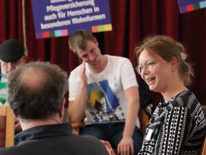 Eine Frau spricht mit einem Mann. Personen sitzen  im Kreis. Die Frau hat braune lange Haare, die sie mit einer Haarspange hochgesteckt hat. Sie trägt eine Brille.