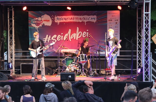 Band auf der Bühne. Gitarrist, Bassist und Schlagzeuger. Publikum vor der Bühne.