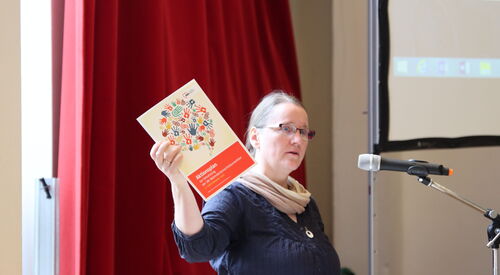 Rosalind Honig bei der Eröffnungsrede des Fachtags. Sie hält dabei ein Exemplar des Aktionplans des AWO Bezirksverbandes mit dem echten Arm und zeigt ihn den Teilnehmer*innen der Tagung