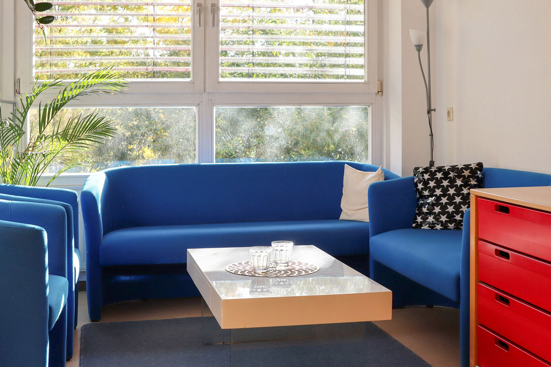 Sprechzimmer. 2 blaue Sessel, 2 Blaue Sofas und ein rechteckiger Tisch in der Mitte.
