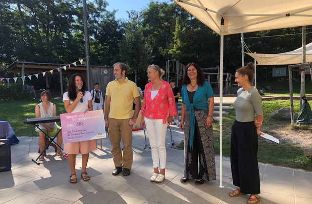 Sonniger Tag mit blauem Himmel. 5 Personen stehen draußen nebeneinder. Frau mit langen schwarzen Haaren, weißem T-Shirt und kurzer Hose hält einen Spendencheck und ein Mikro in der Hand. Die anderen Personen in sommerlicher Kleidung lächeln fröhlich.