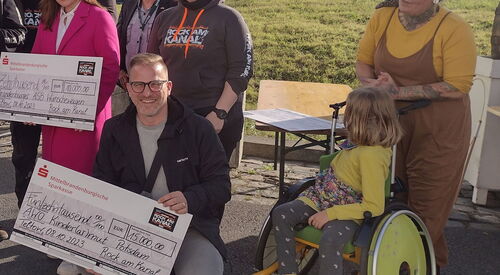 Zwei Spendenschecks werden in die Kamera gehalten. Ein Mann mit Brille, Undercut-Frisur hockt neben Mädchen im gelben Rollstuhl und hält einen Scheck im Wert von 15.000 Euro in der Hand. Dahinter eine Frau im pinken Hosenanzug die den zweiten Scheck hält.