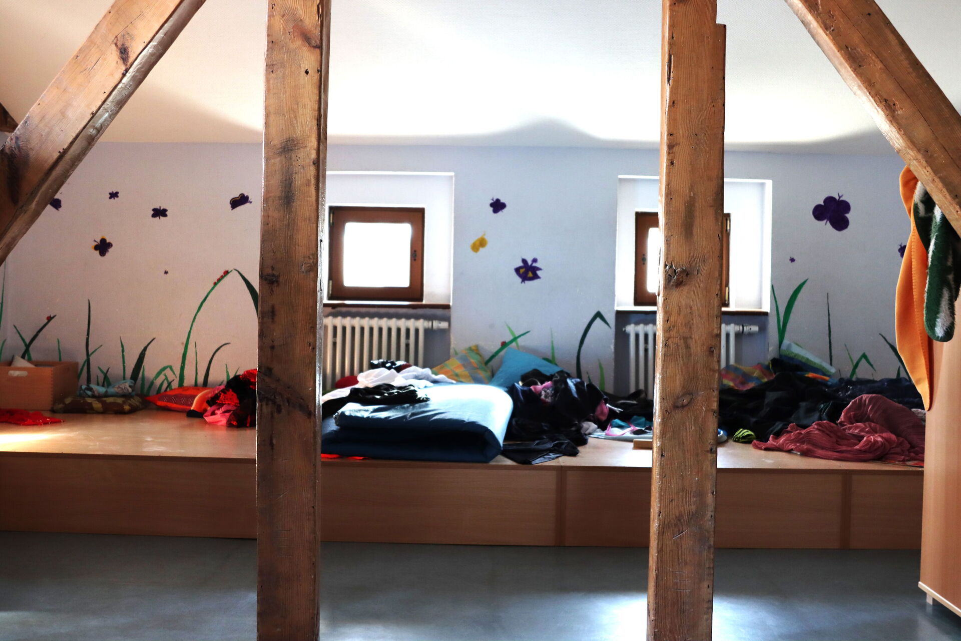 Dachgeschoss. Kleine Fenster. Großer Spielraum der von Holzbalken gestützt wird. Auf einer erhöhten Holzfläche liegen Decken, Kissen und kleidung chaotisch durcheinander.