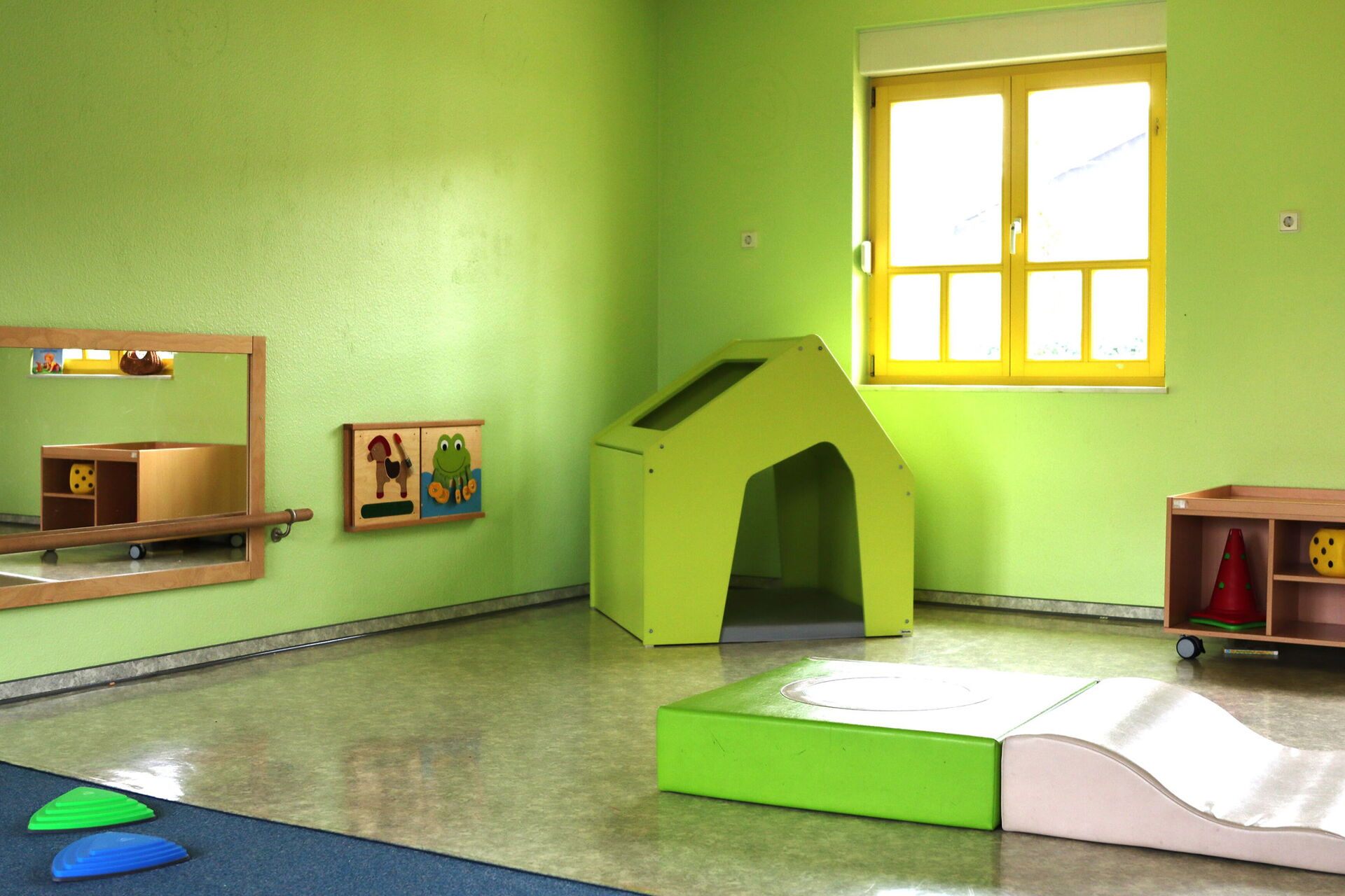 Spielzimmer. Raum mit grünen Wänden. An der Wans ein Spiegel und zwei Bilder, ein Pferd und ein Frosch. In der Ecke steht ein kleines grünes Häuschen mit einer Matte darin. Ein Fenster mit gelben Rahmen durch das die Sonne scheint.