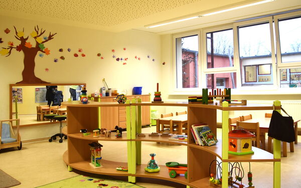 Spielzimmer. Geschwungenes Regal mit Spielsachen für Kleinkinder. An der Wand hinten ein rechteckiger Spiegel. Darüber an der Wand ein kahler Baum mit bunten Herbstblättern aus Papier.