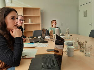 Großer Raum, großer Tisch, 2 Frauen mit Laptops vor sich und eine Frau mit Stift und Notizheft. Auf dem Tisch, Tassen, Teller, Wasserkaraffe und Zettel und Stifte.