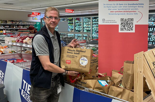 Ein Mann in einem Rewe Markt mit einer gefüllten Papiertüte in den Händen. Es stehen schon mehrere solcher Tüten in einem dafür vorgesehenen Verkaufsständer.