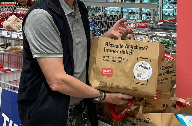 Ein Mann in einem Rewe Markt mit einer gefüllten Papiertüte in den Händen. Es stehen schon mehrere solcher Tüten in einem dafür vorgesehenen Verkaufsständer.