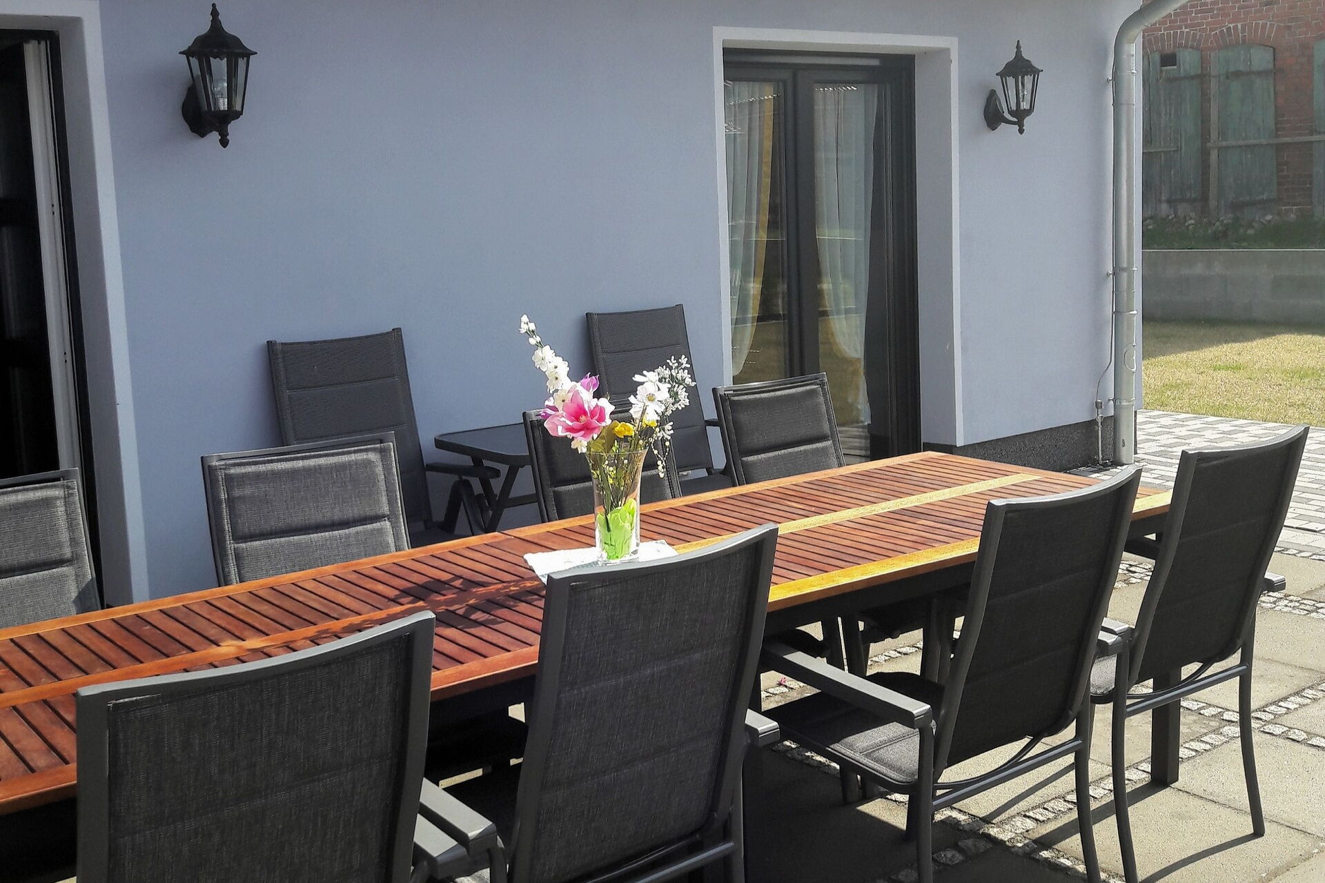 Große Terrasse mit langen Tisch und Stühlen vor einem Haus. Auf dem Tisch steht eine Vase mit Blumen. Die Sonne scheint.