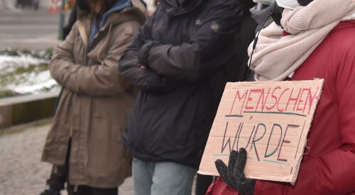 Draußen. Winter. Menschen mit FFP2 Masken in warmer Kleidung. Ein Mensch mit roter Jacke, schwarzen Handschuhen, und hellem Schal hält ein Pappschild vor die Brust. TextMENSCHENWÜRDE