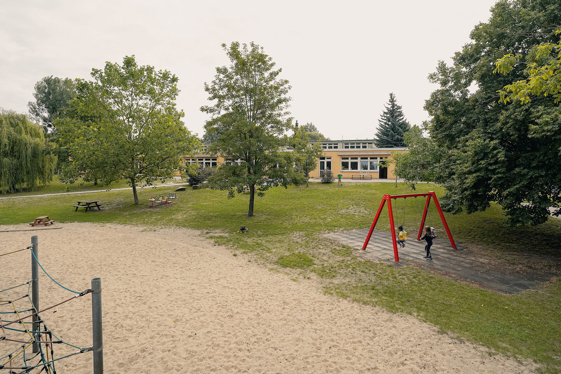 Großes Gelände. Wiese, Bäume, großer Sandkasten. Kinder auf einer roten Schaukel. Spielplatz
