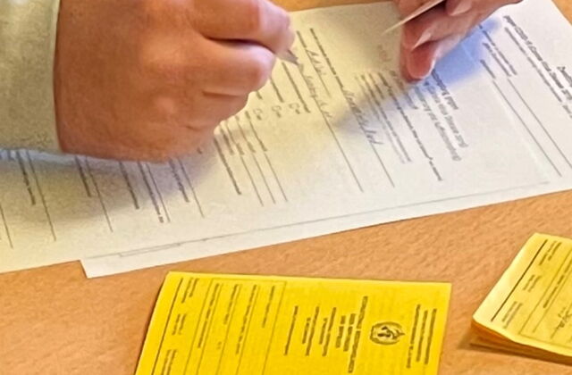 Ein Impfausweis liegt auf dem Tisch. Daneben ein Formular das von einem Mensch mit grauem Pullover ausgefüllt wir. In der linken Hand hält er ein Chipkarte in der rechen einen blauen Kugelschreiber.