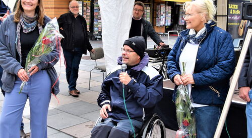 Zwei Frauen mit Blumen in den Händen. Zwischen ihnen ein Rollstuhlfahrer der in ein Mikro spricht. Dahinter ist ein weißer Pavillon. Der Rollstuhlfahrer trägt eine schwarze Mütze, Brille, Regenjacke. Er hat einen Aktivrollstuhl.