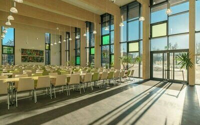Großer Saal, mit vielen Tischen und Stühlen. Große Fenster die viel Licht herein lassen. Hinten ist eine große Glastür die Sonne scheint in den Raum.