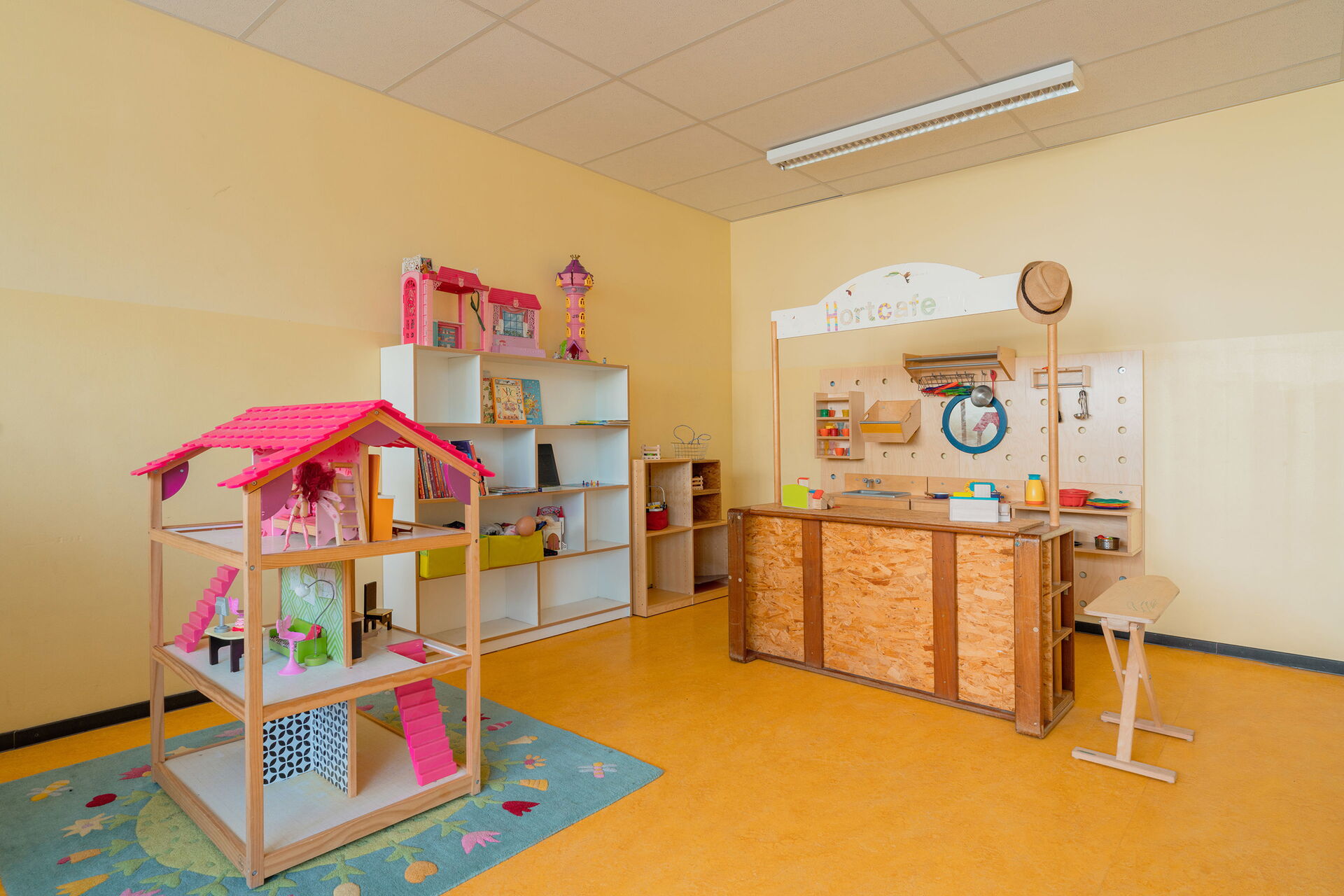 Spielzimmer. Puppenhaus mit pinken Dach auch einen geblümten kleinen Kinderteppich, Regal an der Wand mit Spielzeug. Und ein Spielzeug "Hort-Café" aus Holz mit Tresen und Ablagen für Geschirr dahinter an der Wand.