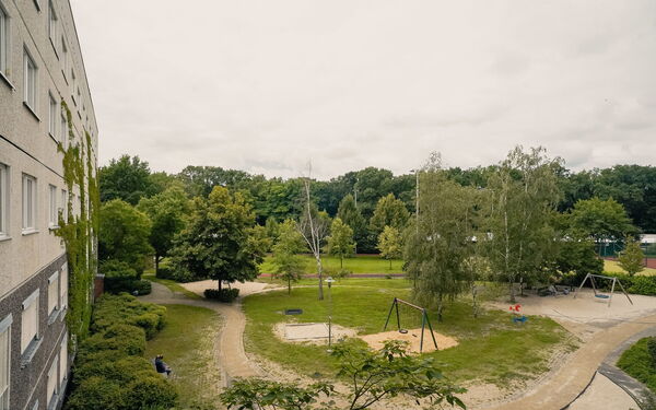 Blick aus einem oberen Fenster nach draußen auf einen Spielplatz. Sandkasten, Schaukeln, Wiese, ein paar Bäume.