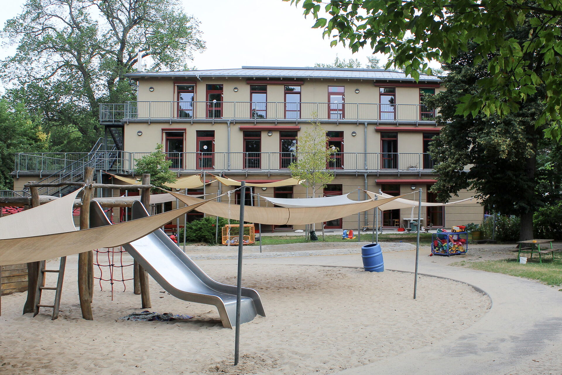 Spielplatz mit großen Sandkasten, Klettergerüst mit Rutsche. Im Hintergrund das dreistöckige Kitagebäude.