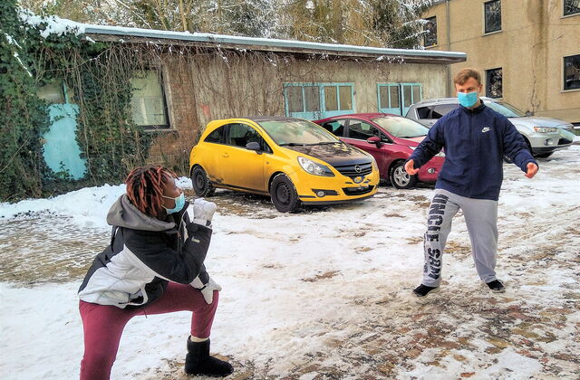 Tanzszene im Freien. Es ist Winter, Schnee liegt, parkende Autos. Eine schwarze Tänzerin mit OP Maske ist in einer breitbeinigen Hocke einem Tänzer mit OP Maske zugewandt. Der weiße Tänzer hat die Arme ausgebreitet. Sie stehen etwas von einander entfernt.