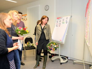 Frau mit Blumenstrauß steht vor einem Flipchart. Eine weitere Frau hat einen Bumenstrauß in der Hand. Die 4 Personen schauen interessier auf eine Tafel mit Notizen.