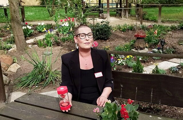 Frau sitzt an einem Tisch im Garten, Blumen blühen in den Beeten. Die Frau hat sehr kurze sunkle Haase, eine große runde, schwarz gerahmte Brille, sie trägt einen schwarzen Blazer über ein schwarzes Oberteil. Sie hält ein rote Sanduhr in der rechten Hand.