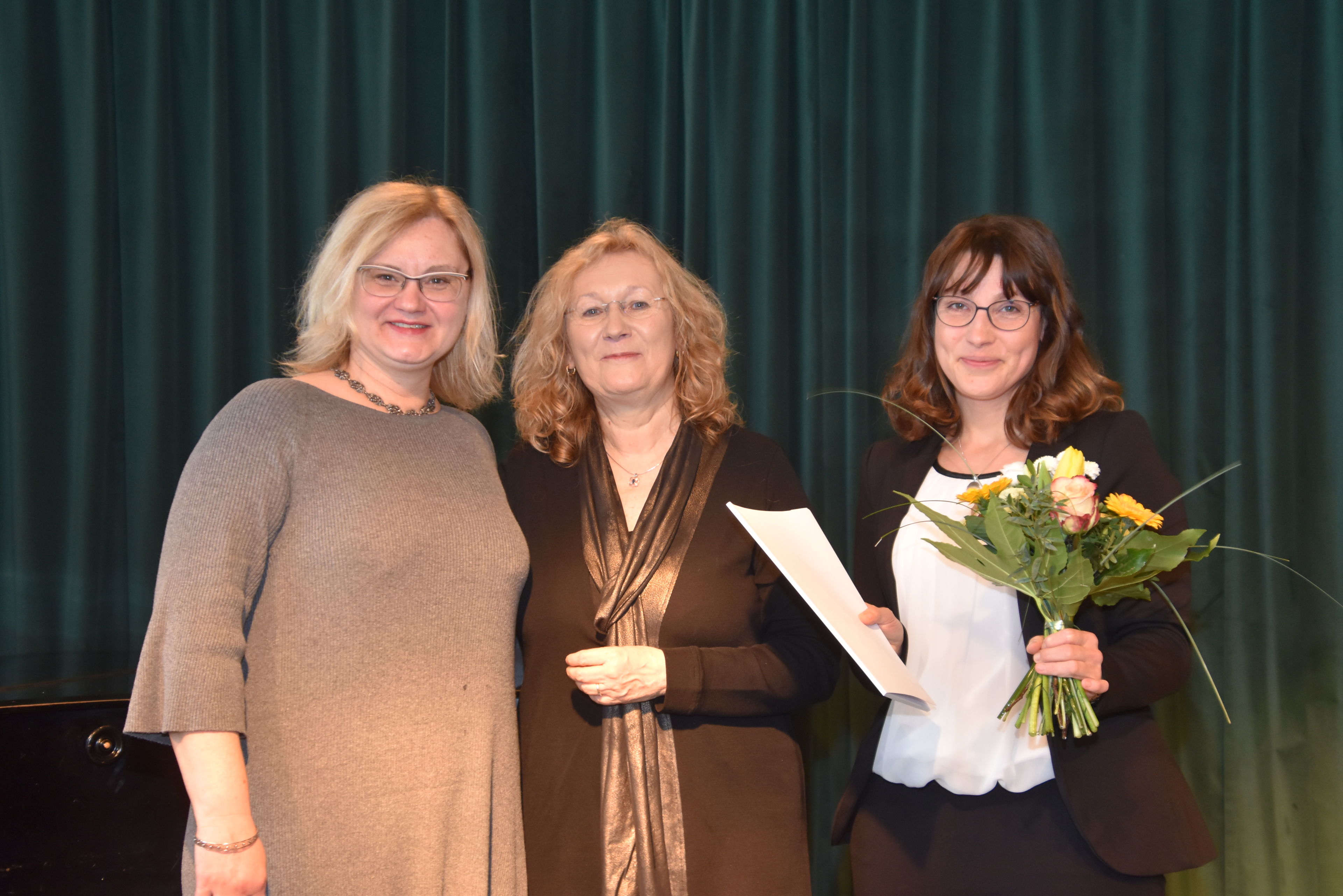 Drei Frauen vor dunkelgrünem Vorhand. Eine Frau mit brauen langen Haaren, Brille, Blumen und Zeugnis in der Hand, lächelt in die Kamera.