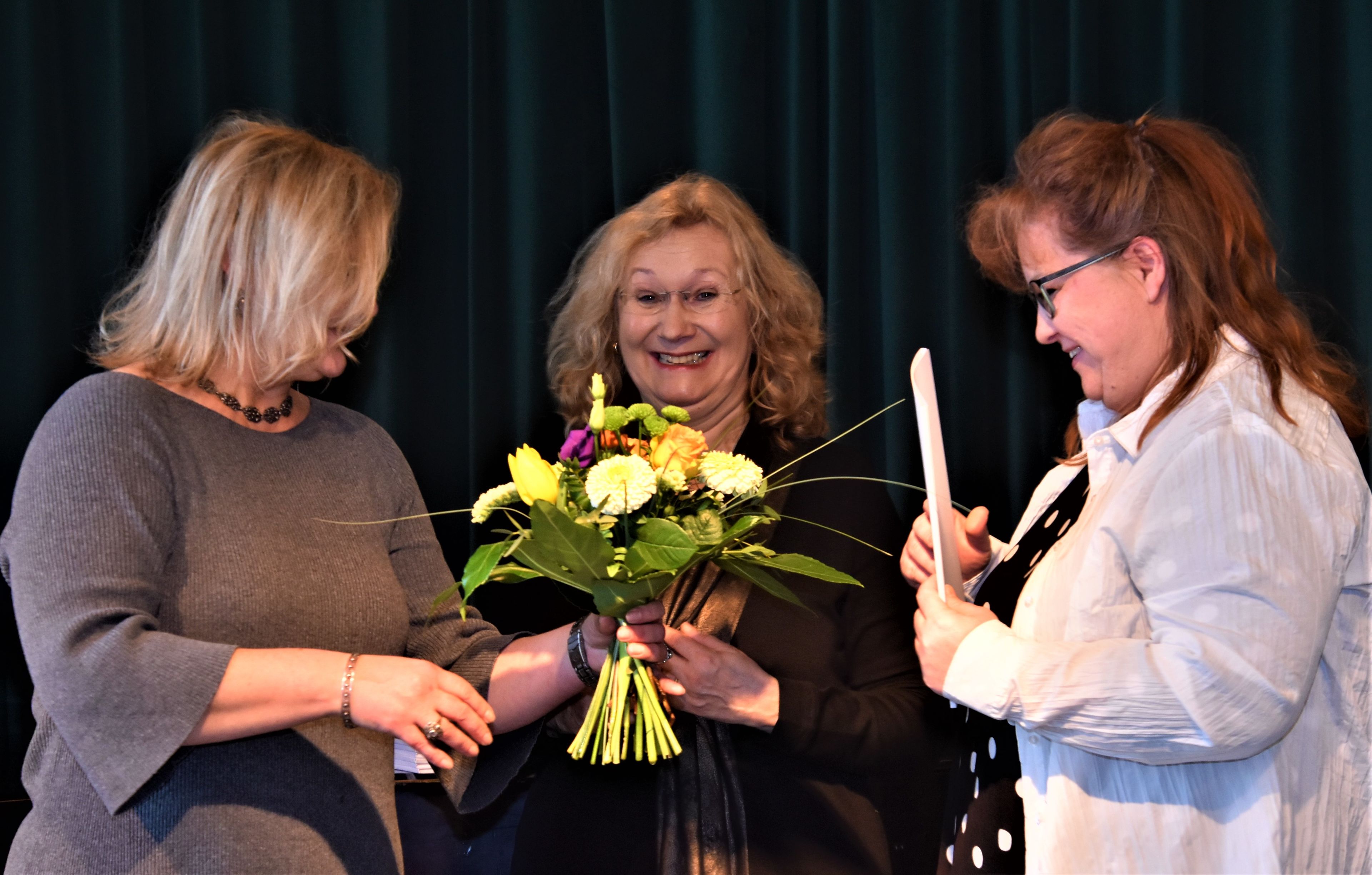 Drei Frauen vor dunkelgrünem Vorhand. Blumenstrauß und Zeugnis werden an weibliche Schulgesundheitsfachkraft übergeben. Glückliche Gesichter.