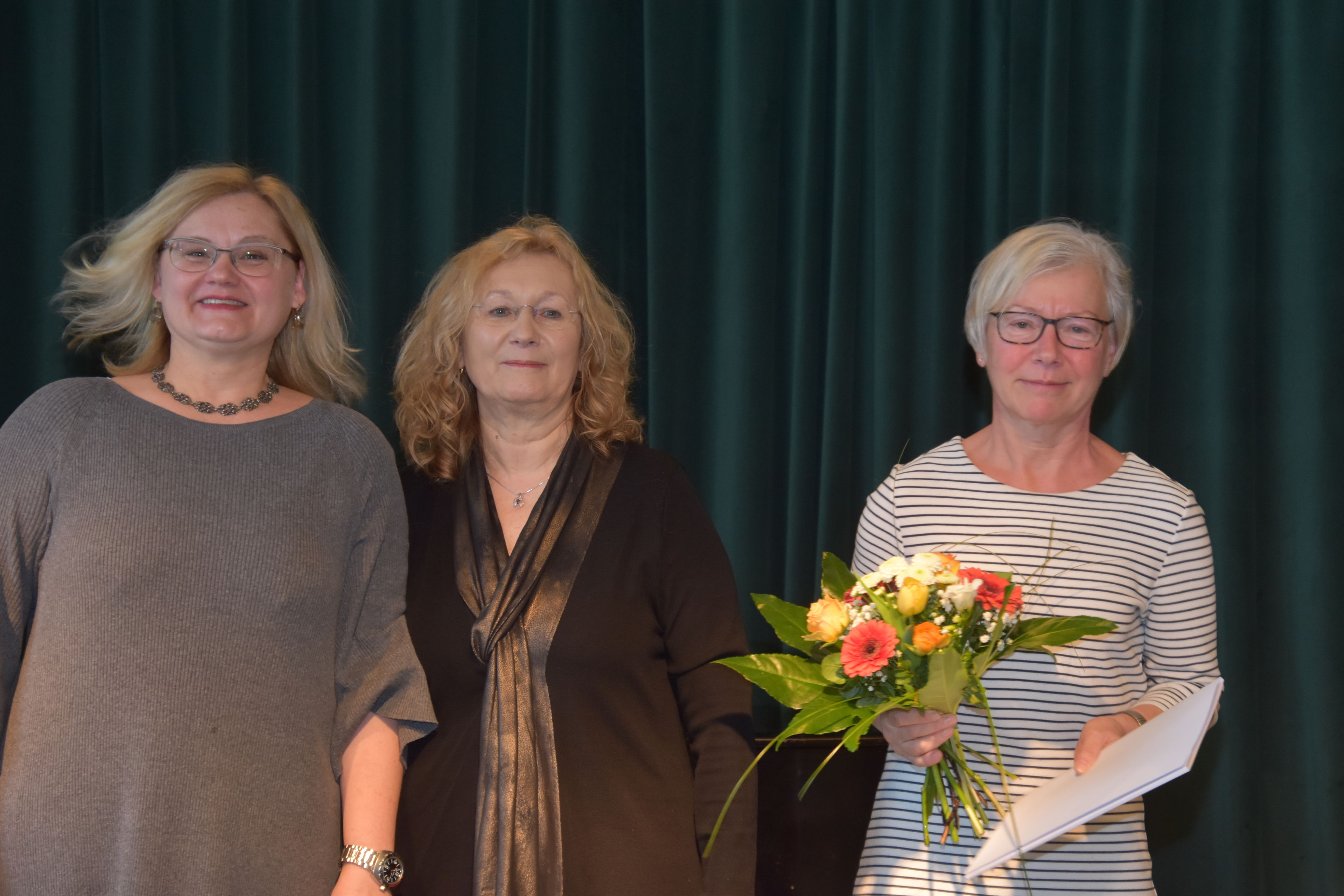 Drei Frauen vor dunkelgrünem Vorhand. Eine ältere Frau mit Blumenstrauß und Zeugnis in den Händen. Glückliche Gesichter.